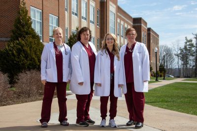 A group photo of the department of nursing faculty wearing maroon scrubs and white coats near Concord University's bell tower
